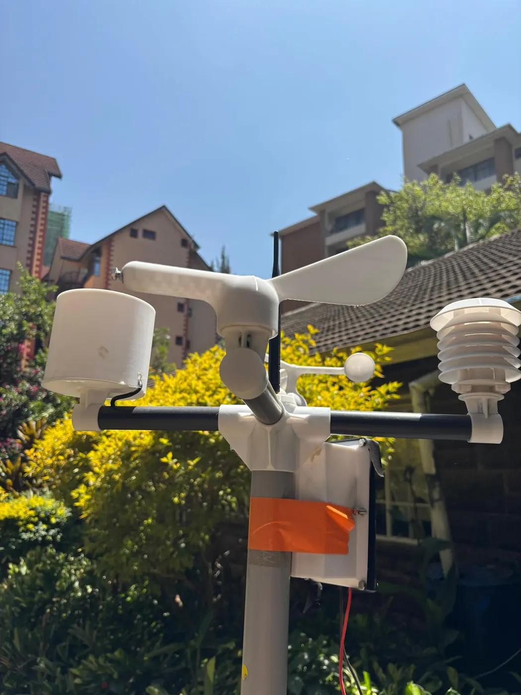 Weather station head with wind vane, spinning cups, rain gauge and sensor shield against blue sky over garden shrubs.